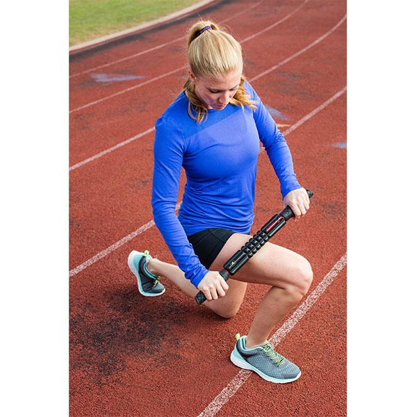 "A woman kneels on a running track to use the TriggerPoint GRID STK® X Foam Roller Massage Tool on her thigh."